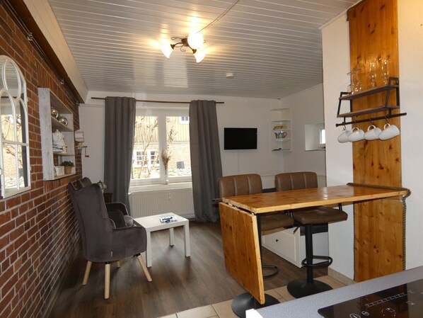 A compact room with a dining table, two chairs, a small white table, a TV mounted on the wall, and a window with curtains.