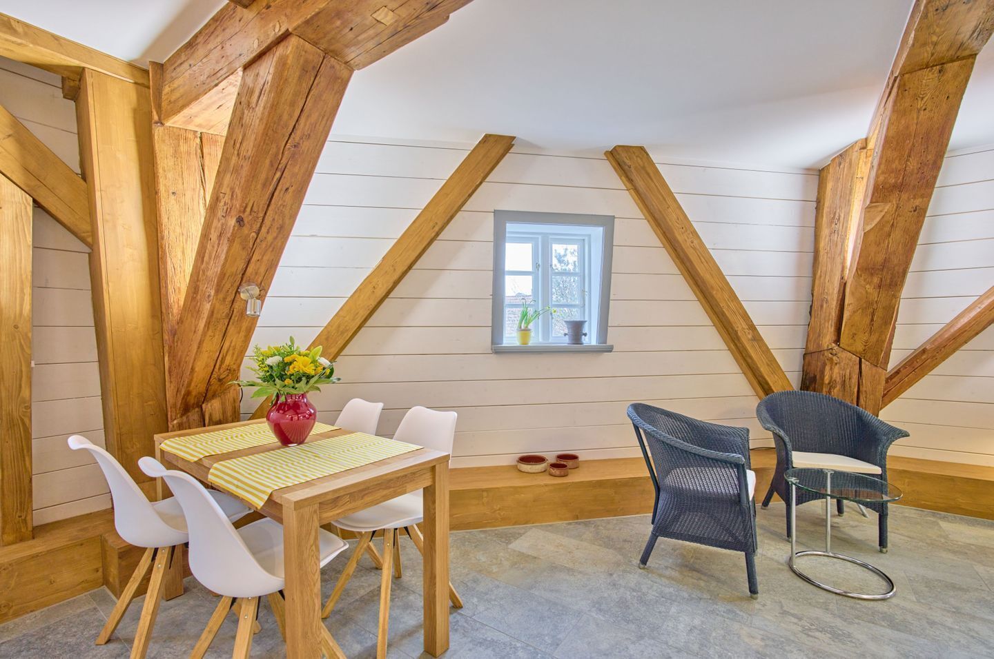 Ein Zimmer mit geneigter Decke, Holzbalken, einem Esstisch mit Stühlen, einer Vase mit Blumen und einem Fenster mit Aussicht.