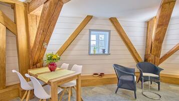 Ein Zimmer mit geneigter Decke, Holzbalken, einem Esstisch mit Stühlen, einer Vase mit Blumen und einem Fenster mit Aussicht.