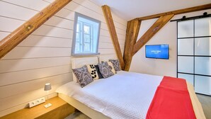Un dormitorio con techo inclinado, una ventana, cama con almohadas estampadas, mesita de noche con lámpara y televisor montado en la pared.