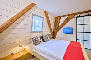 Ein Schlafzimmer mit schrägem Dach, einem Fenster, einem Bett mit gemusterten Kissen, einem Nachttisch mit Lampe und einem an der Wand montierten Fernseher.