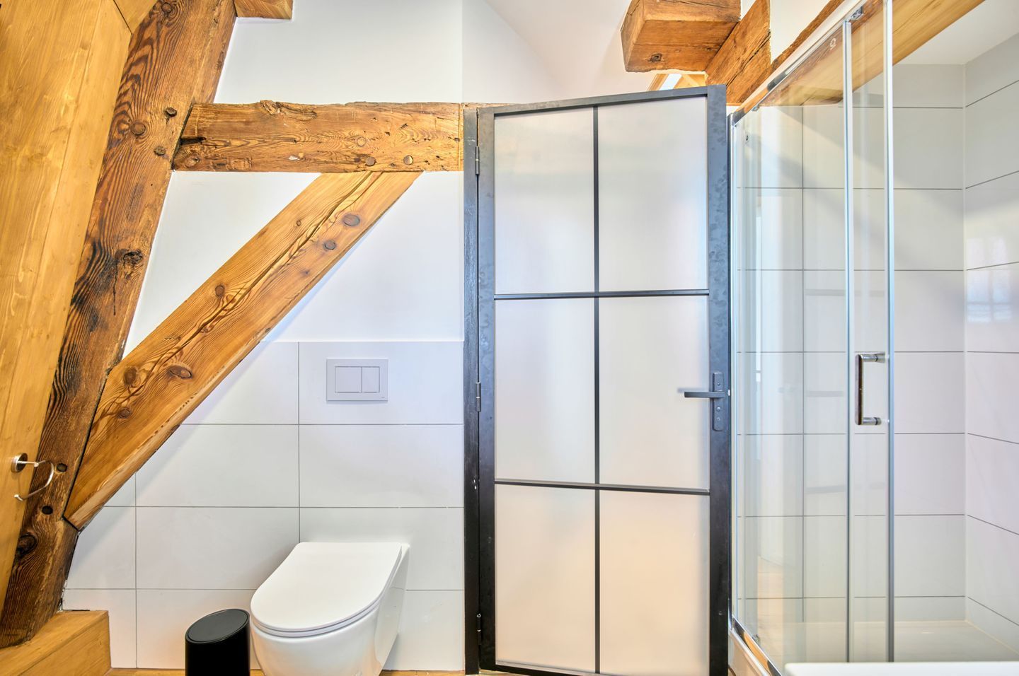 Un baño con techo de madera, inodoro blanco, cabina de ducha con mampara de cristal y pared de azulejos blancos.
