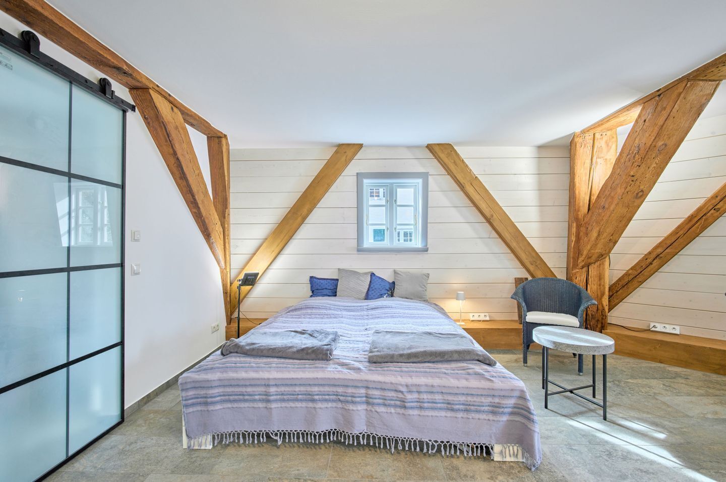 Ein Schlafzimmer mit Bett, Stuhl, Fenster und Holzbalken.