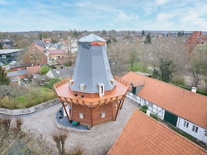 Ein hölzernes Turmhaus mit einem kegelförmigen Dach, umgeben von einem Kieshof, neben traditionellen Häusern.