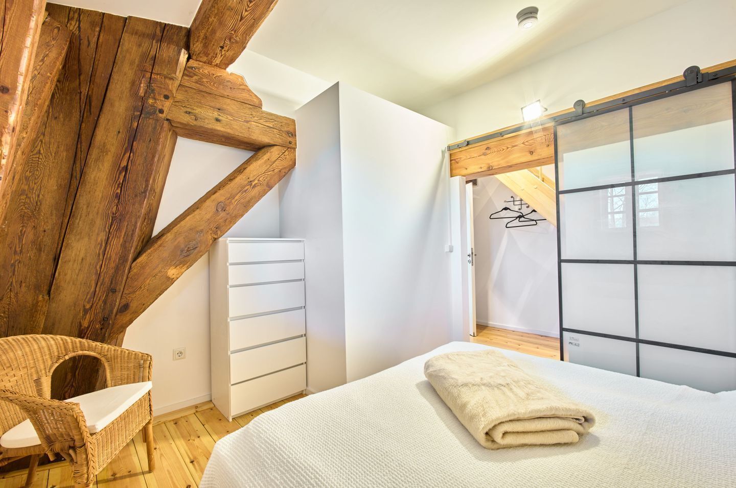 Une chambre à coucher avec un lit, une commode blanche, une chaise en osier et une structure de plafond en bois.