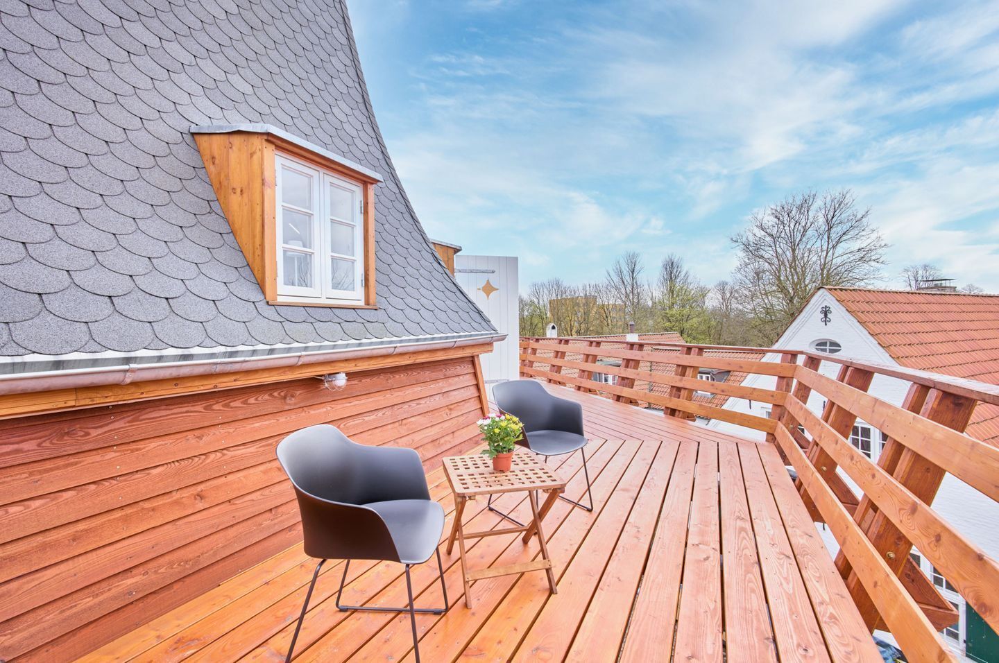 Eine Holzterrasse mit zwei Stühlen, einem Tisch, einem Fenster und einem geneigten Dach.