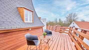 Eine Holzterrasse mit zwei Stühlen, einem Tisch, einem Fenster und einem geneigten Dach.