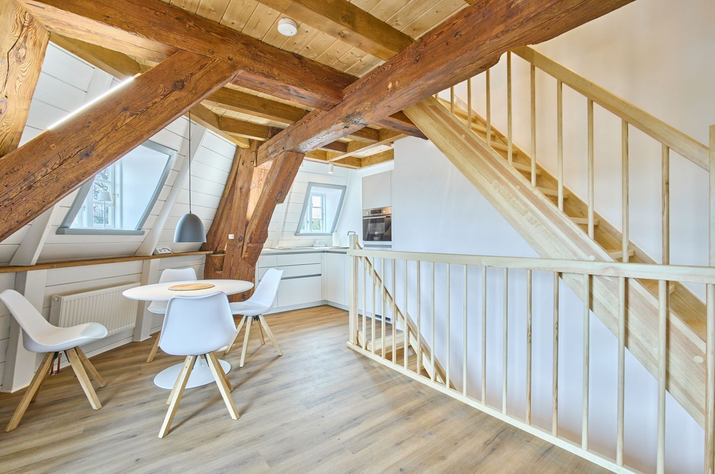 Ein Loft-Zimmer mit Holzboden, einer Essecke mit weißen Stühlen, einer Küche mit weißen Schränken und einer Holztreppe.