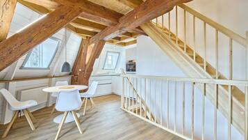 Ein Loft-Zimmer mit Holzboden, einer Essecke mit weißen Stühlen, einer Küche mit weißen Schränken und einer Holztreppe.