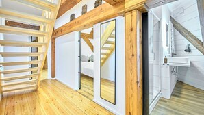 Una habitación con piso de madera, una escalera, una pared blanca y un baño con lavamanos y espejo.