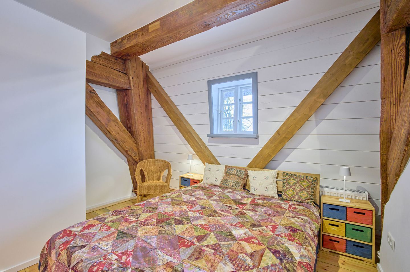 Ein Schlafzimmer mit Holzboden, einem Bett mit bunter Steppdecke, einem Korbstuhl und einem Fenster.