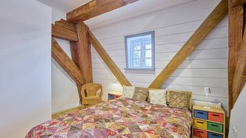 Ein Schlafzimmer mit Holzboden, einem Bett mit bunter Steppdecke, einem Korbstuhl und einem Fenster.