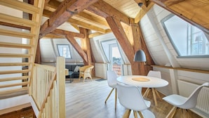 A loft-style room with exposed wooden beams, a round table with chairs, a sofa, and a window.