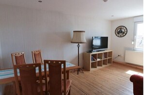 Ein Wohnzimmer mit Holzboden, einem Esstisch mit Stühlen, einem Fernseher auf einem Schrank, einer Lampe und einer Wanduhr.