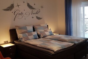 Un dormitorio con cama, dos mesitas de noche, un adorno en la pared y una ventana con cortinas.