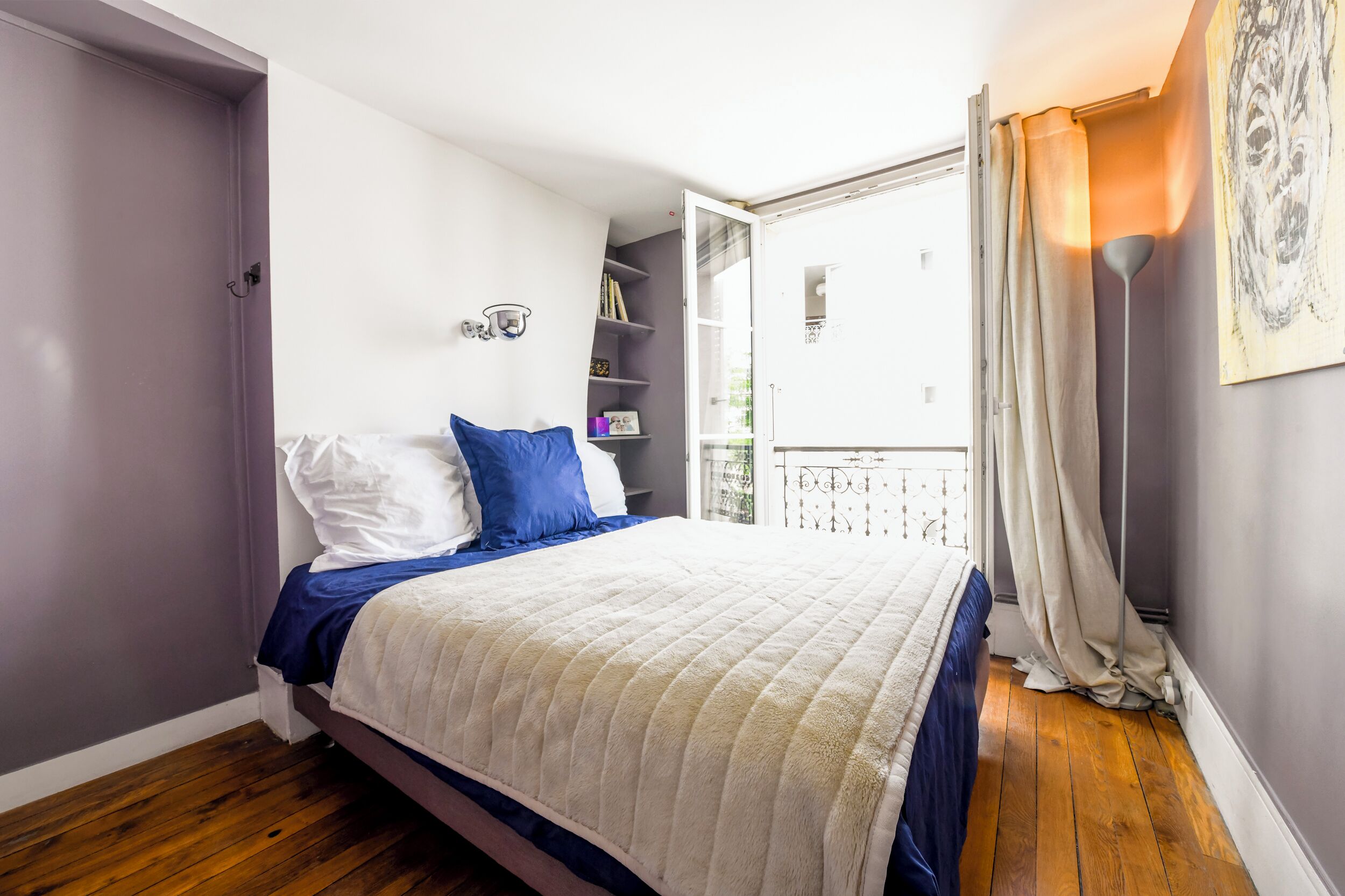 Un dormitorio con cama, armario, estantería con libros y balcón con cortinas.