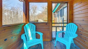 Una cabaña de madera con dos sillas Adirondack, una terraza de madera y vistas a un bosque a través de ventanas amplias.