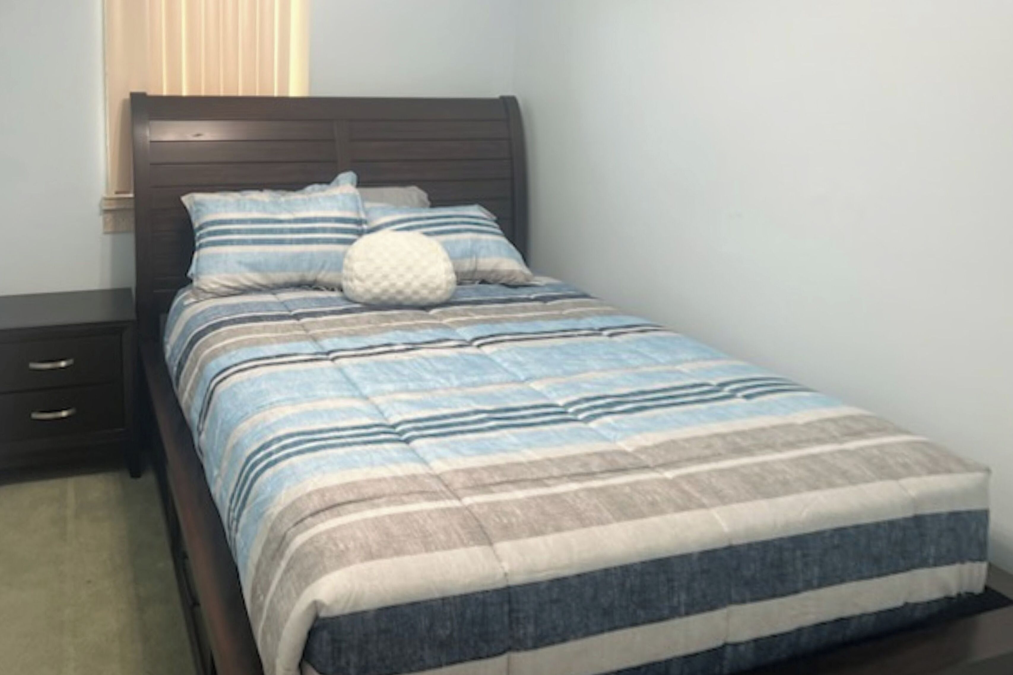 Un letto con copriletto a righe, una testiera in legno e un comodino con cassetti.