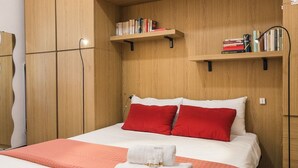 Siististi järjestetty hotellihuone, jossa on sänky, punaiset ja valkoiset tyynyt, valkoinen peitto, punainen huopa sekä kaksi hyllyä, joilla on kirjoja ja lamppu.