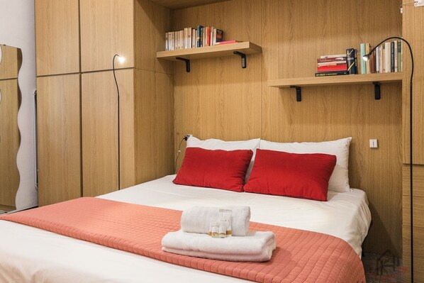Ein ordentlich eingerichtetes Hotelzimmer mit einem Bett, roten und weißen Kissen, einer weißen Decke, einer roten Tagesdecke sowie zwei Regalen mit Büchern und einer Lampe.