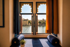 A room with a view of a river and buildings, featuring a window with a decorative frame, a bed with a blue bedspread, and a potted plant. - Hotel The Nayee Haveli - Lake View Hotel (Udaipur)