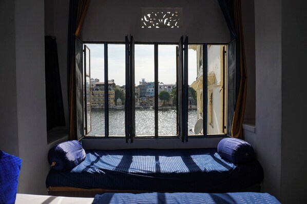 A room with a bed, a window with curtains, and a view of a river and buildings. - Hotel The Nayee Haveli - Lake View Hotel (Udaipur)