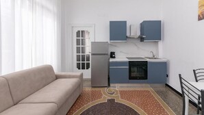 A compact kitchen with a refrigerator, stove, and sink, a beige sofa, and a patterned rug. - GALIA ROOMS (Genoa)