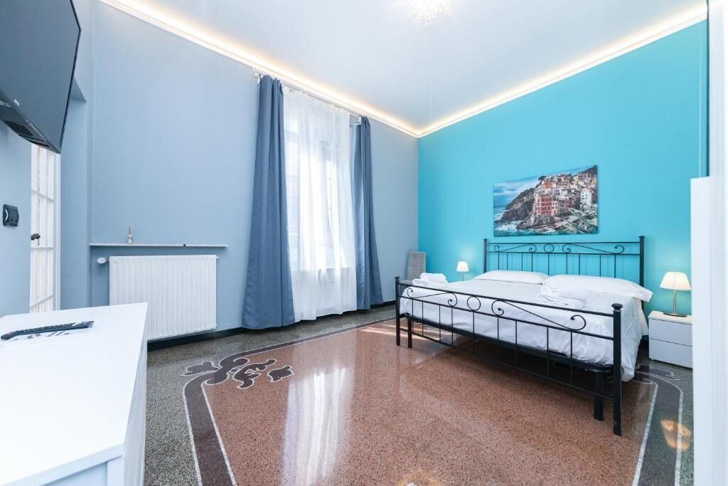 A bedroom with a bed, a desk, a radiator, curtains, a painting, and a lamp.