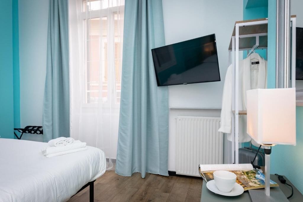 A hotel room with a bed, a TV mounted on the wall, a bedside table with a cup and a book, and a window with curtains.