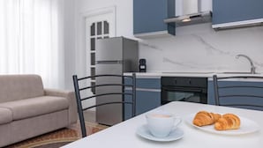 A modern kitchen with a refrigerator, stove, and coffee maker, a dining table with croissants, and a sofa. - GALIA ROOMS (Genoa)