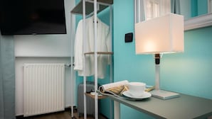 A hotel room with a TV mounted on the wall, a radiator, a white shelf with a white robe, a table with a lamp, a cup, and a newspaper. - GALIA ROOMS (Genoa)
