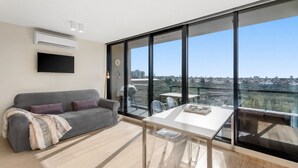 Un salón moderno con un sofá gris, una mesa de vidrio y un ventanal con vistas a la ciudad.