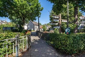 A paved pathway leading to a traditional building with a sloped roof, surrounded by lush greenery and residential houses.