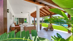 A covered outdoor seating area with wicker furniture, a pool in the background, and lush greenery. - 4 bedroom lovely home in Roche (Roche)