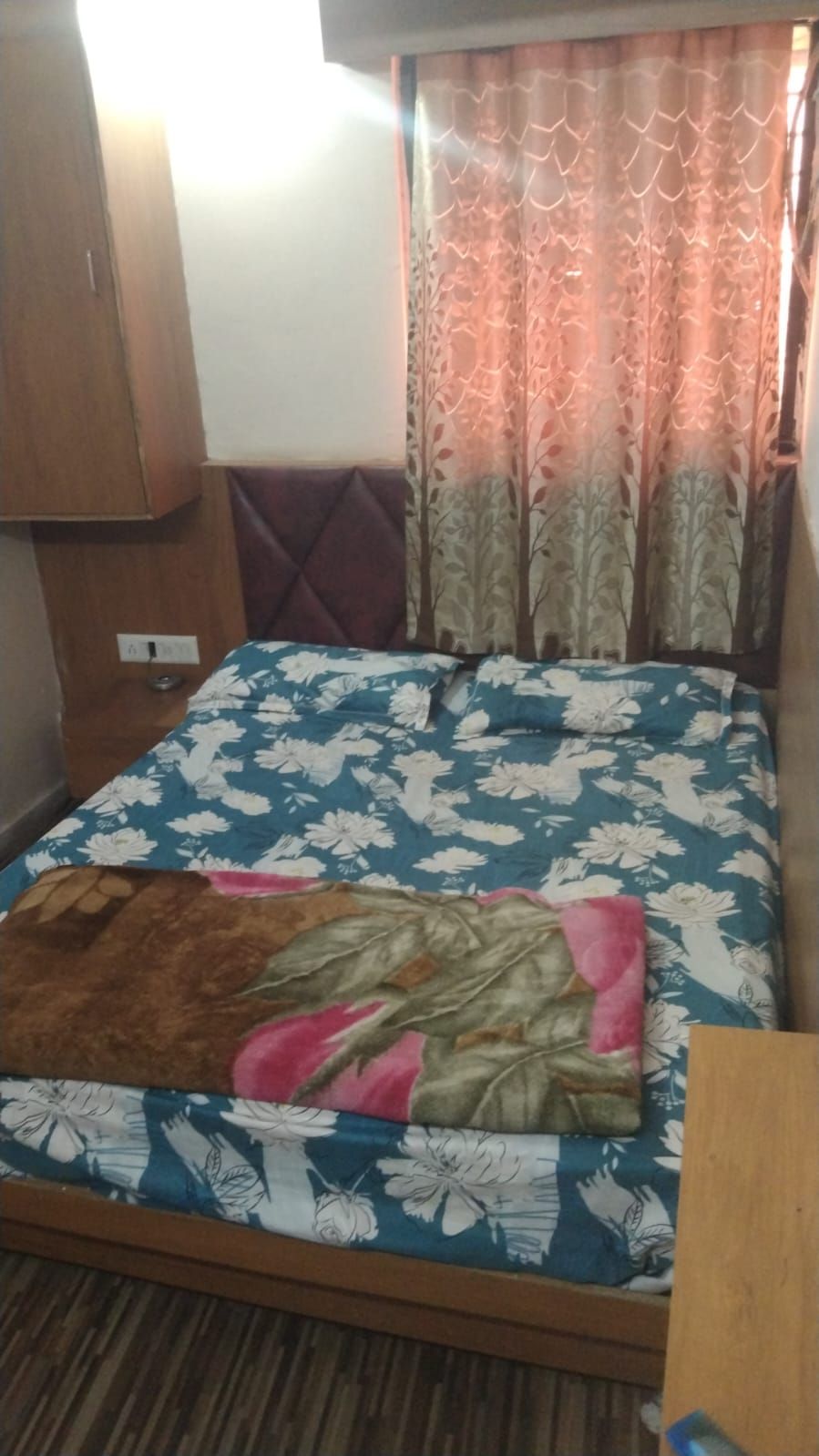 A bed with a floral bedspread, a wooden headboard, and a window with patterned curtains.