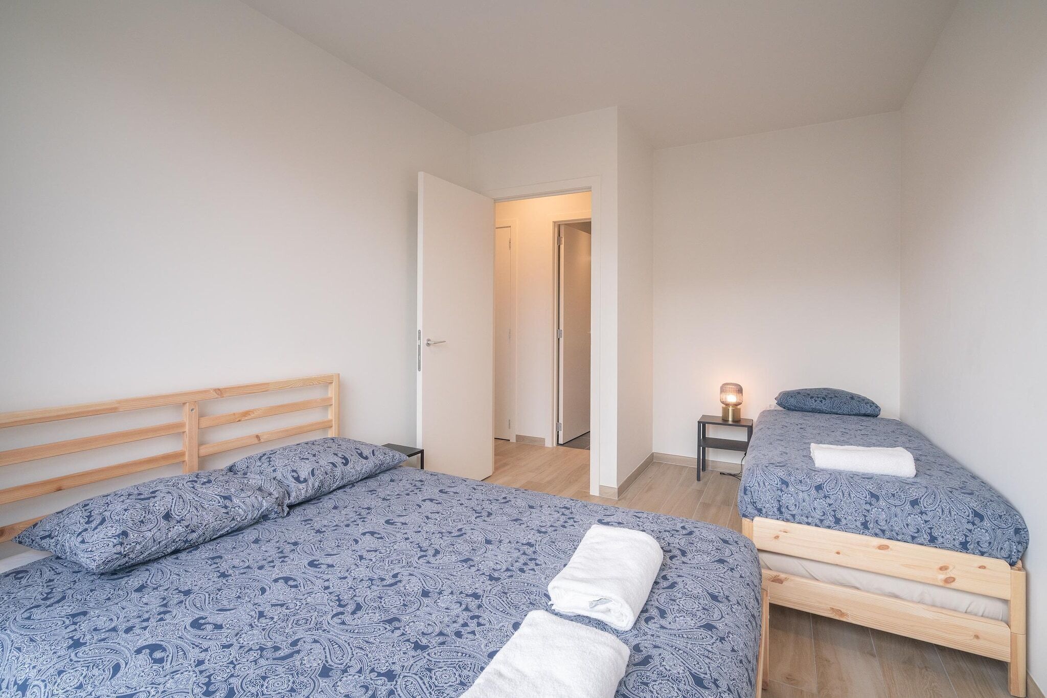 A bedroom with two beds, a wooden headboard, a bedside table with a lamp, and a door leading to another room.