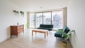 A spacious living room with a wooden coffee table, a green sofa, a wooden dresser, and large windows offering a view of buildings. - Stunning Central Antwerp Stay for 12 (Antwerp)