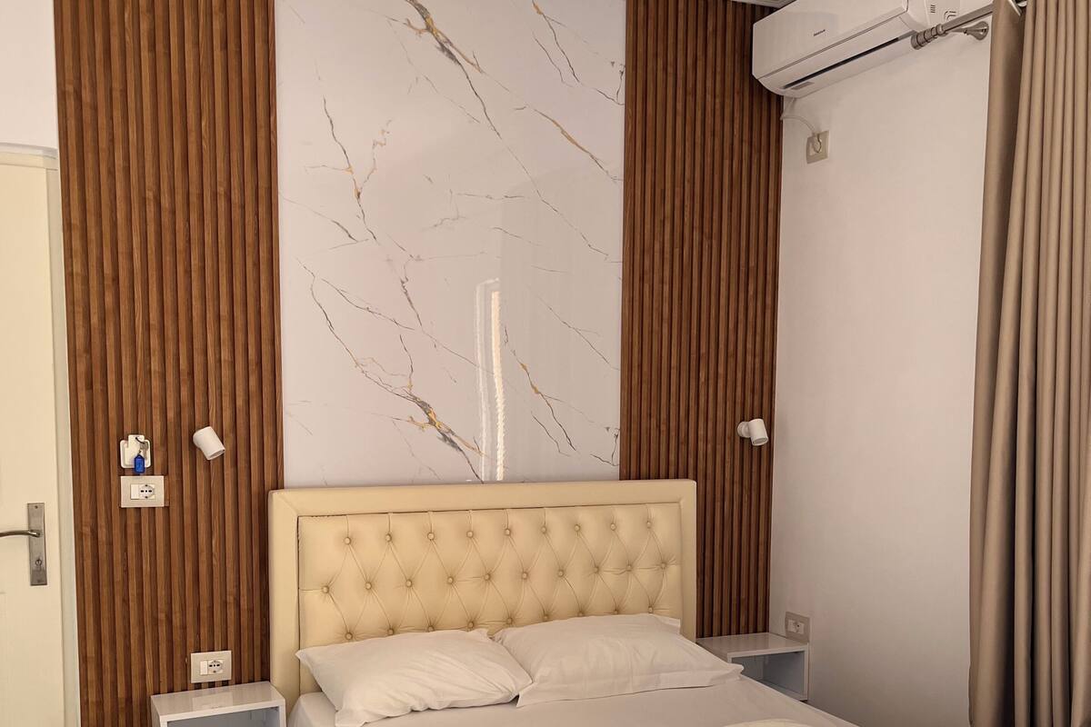 A hotel room with a marble-patterned wall, a tufted headboard, a bed with white linens, a bedside table, and an air conditioning unit.