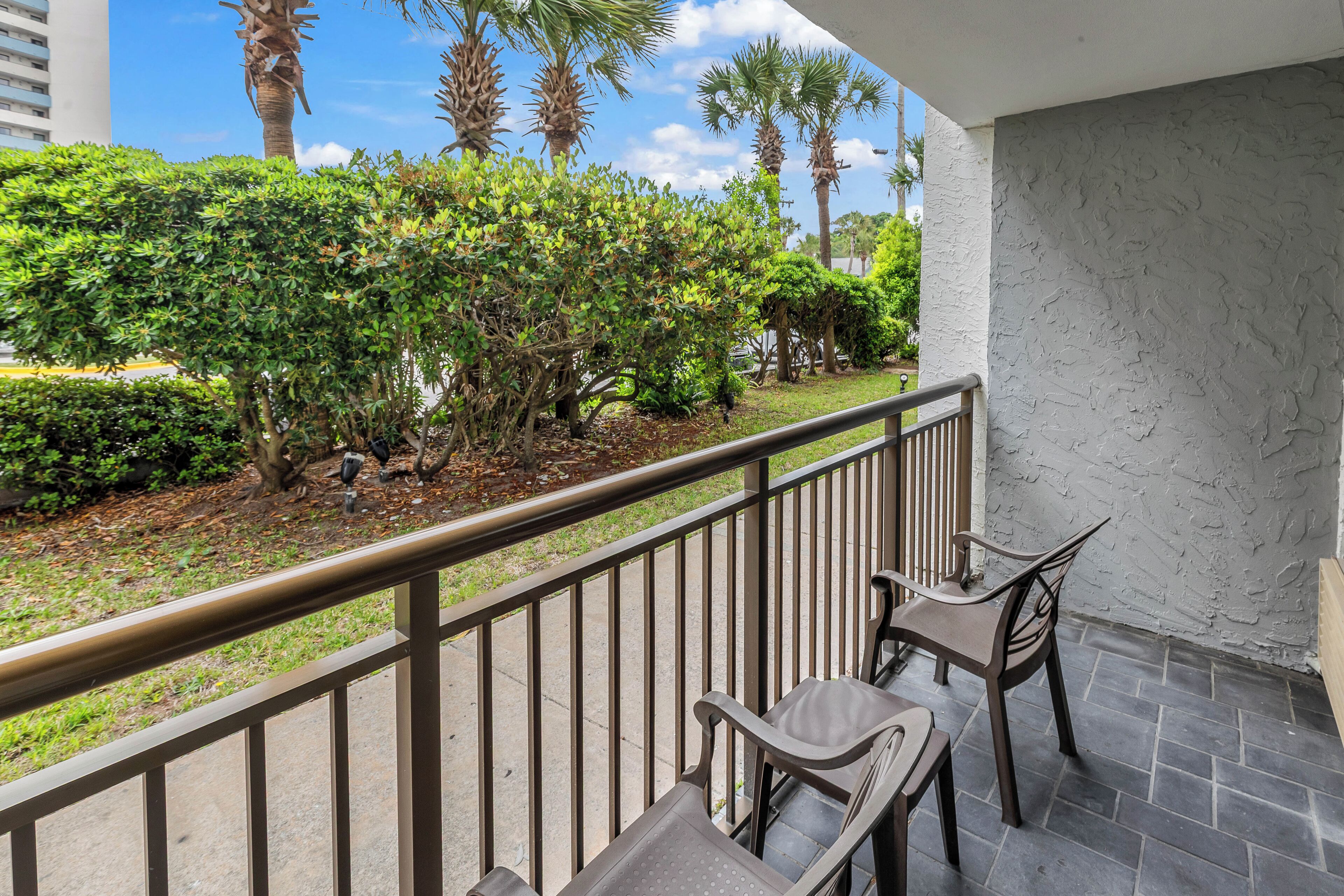 A balcony with a railing, two chairs, and a view of a landscaped garden with trees and shrubs.