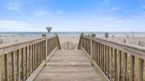 Una passerella in legno che conduce a una spiaggia, con un segnale di avvertimento e un cartello che recita 'PROTEGGI LE DUNE'.