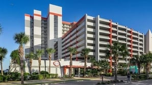 Un hotel a più piani con parcheggio, palme e un cielo azzurro intenso.