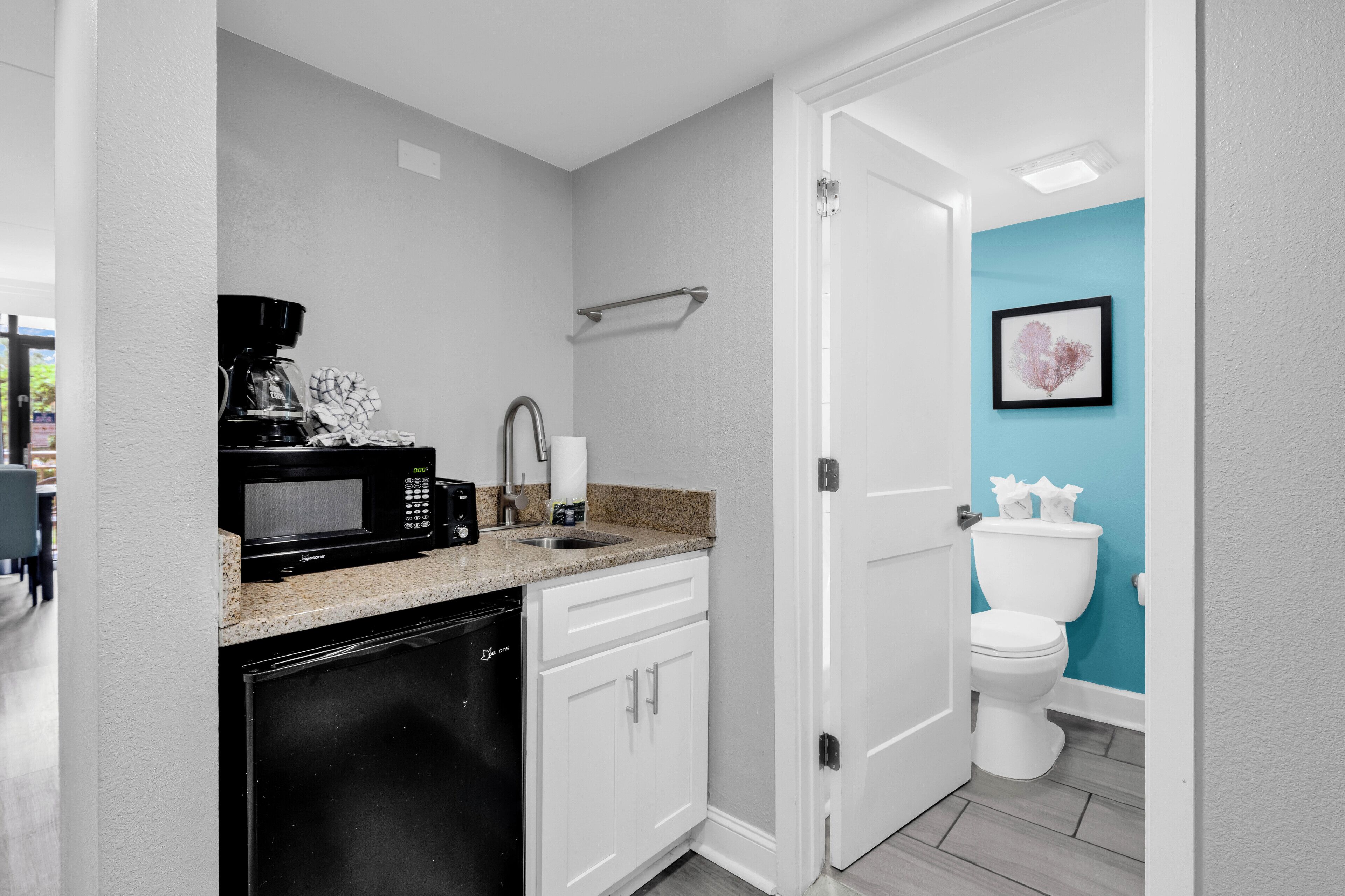 A compact kitchen with a microwave, coffee maker, and dishwasher, adjacent to a bathroom with a toilet and a blue accent wall.