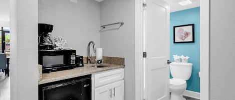 Cocina compacta con microondas, cafetera y lavavajillas, junto a un baño con inodoro y una pared de acento azul.