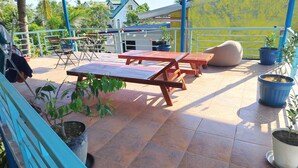 A covered outdoor area with wooden tables and chairs, potted plants, and a view of a house in the background. - Blu Corner - a place to relax (Lian)
