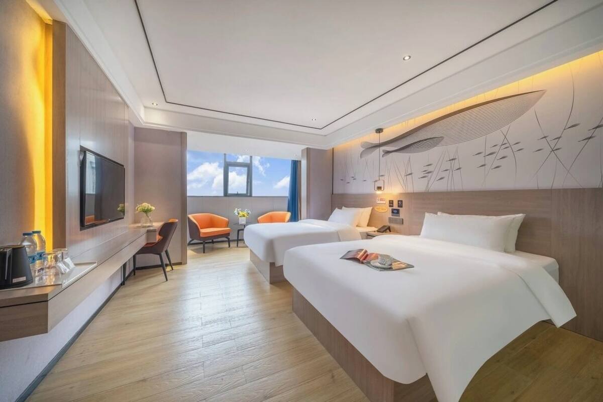 Habitación de hotel con dos camas, un escritorio con televisión de pantalla plana, un pequeño rincón de descanso con sillas y un amplio ventanal con vistas al cielo.