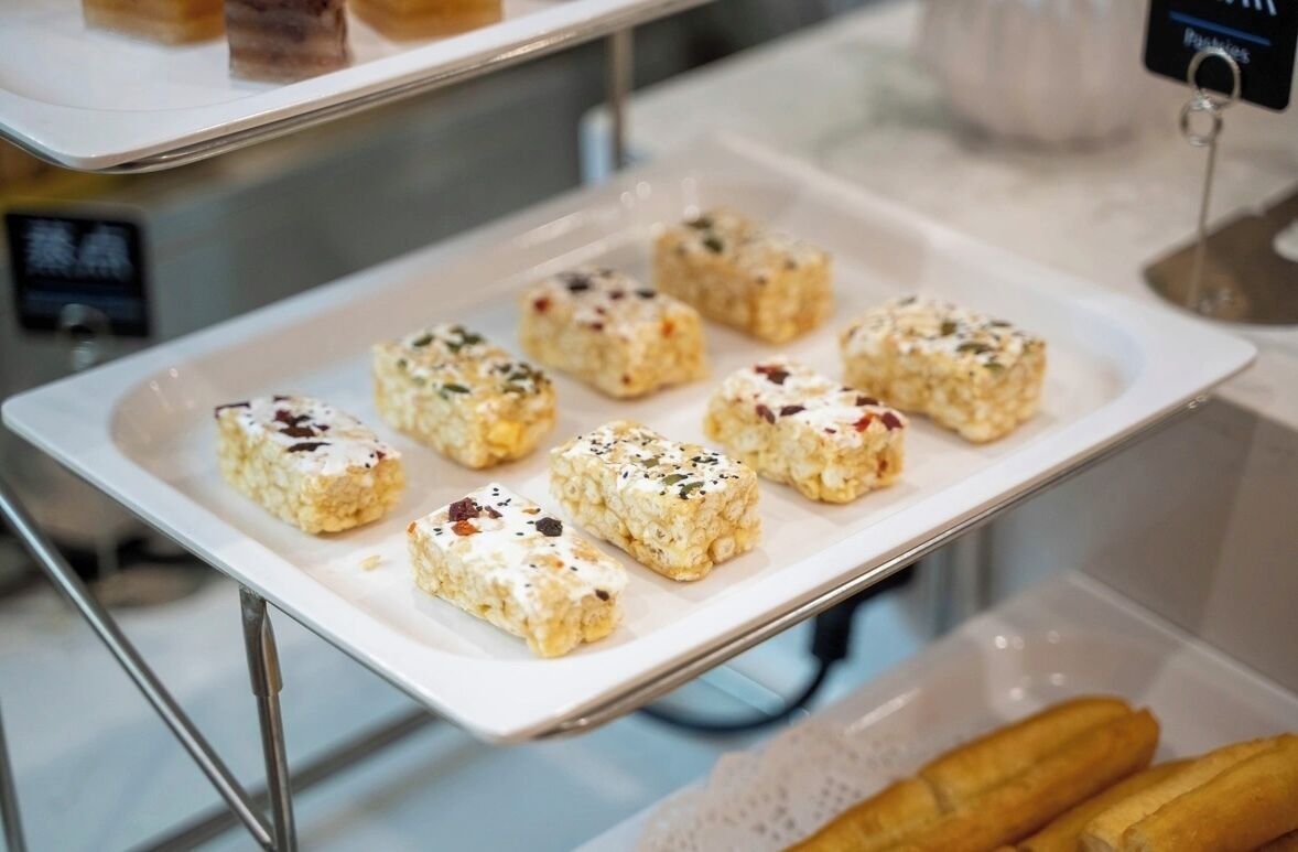A tray of square-shaped food items with seeds on a white plate, a sign with the word 'Pastries,' and a container with baguettes in the background.