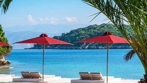 A seaside resort with red umbrellas, lounge chairs, and a view of the ocean and hills. - Bella Mare Hotel (Corfu)