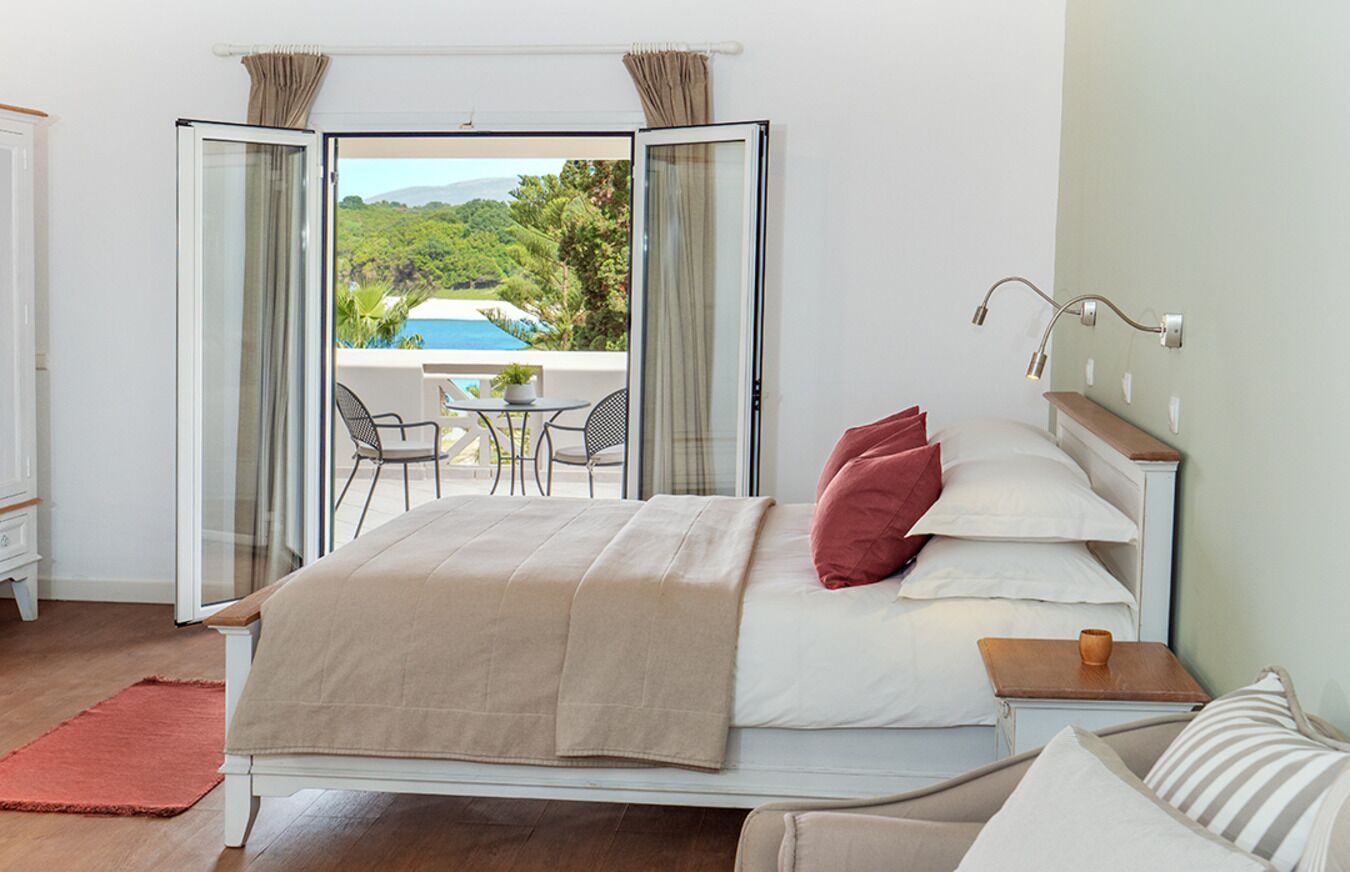 A bedroom with a bed, a bedside table, a chair, and a view of a pool and greenery through an open door.