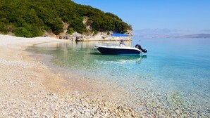 A boat on a pebble beach with clear water and a forested hillside in the background. - Bella Mare Hotel (Corfu)