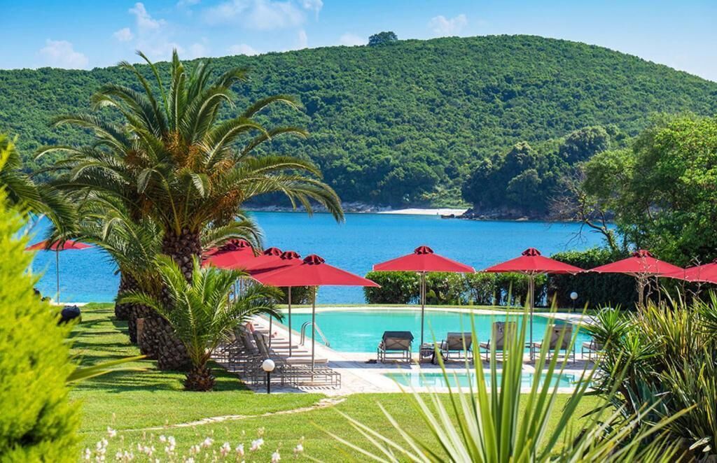 A pool area with red umbrellas, lounge chairs, and a view of the sea and forested hills.